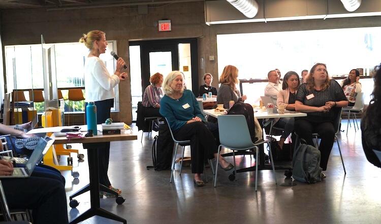 a standing person speaks into a microphone, with an audience seated at 3 roundtables of 6 each and a wall of large windows in the background