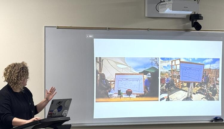 The speaker gestures toward a slide that displays two photos of a map being presented to a group of people outdoors