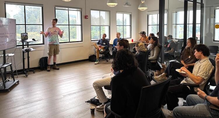 a person stands next to a large TV, gesturing, while 12 people sit and listen to the presentation, in a room with large windows
