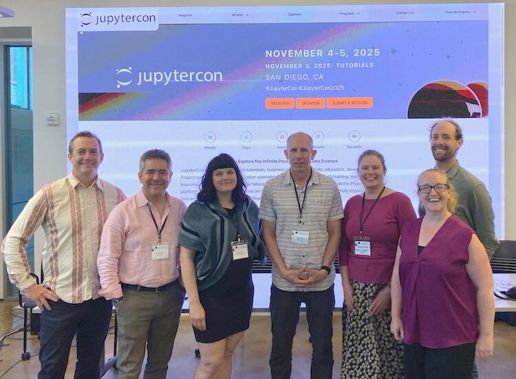 A group of the 6 panelists and 1 moderator standing side by side with a slide projected behind them, featuring "JupyterCon" and the date November 4-5, 2025
