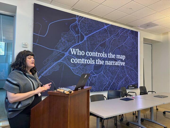 Ciera Martinez stands behind a podium with a projected slide that says "Who controls the map controls the narrative"