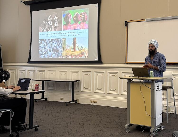 Manvir Singh stands behind a podium in a large room as he presents to a seated audience, with one person and their laptop in the foreground of the photo