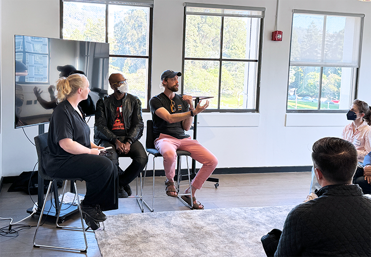  3 people sit on taller chairs with windows behind them in an informal setting, speaking to event attendees